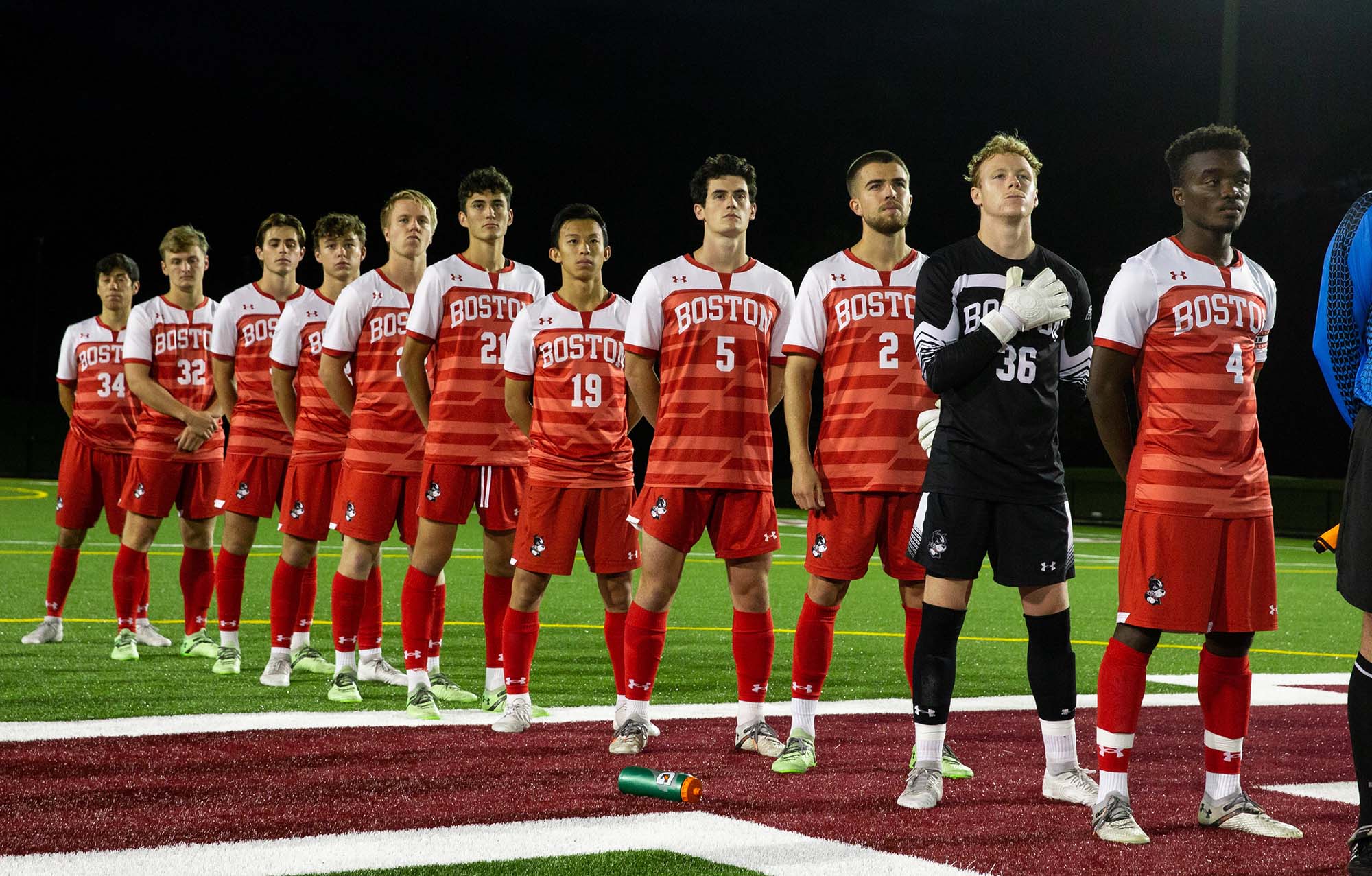 Photo of Kargbo (far right) with his teammates on the field during a fall 2021 game.  They all stand in a line wearing red and white soccer uniforms that read "Boston" as well as their player numbers. The goalie stands at the front in a black and white version of the same uniform.