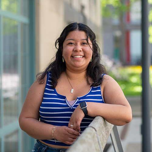Photo of Newbury center grad Cecilia Jarquin Tapia. Tapia wears a striped blue and white tank top and blue jeans and leans and poses against a wooden bench as she smiles to the camera.