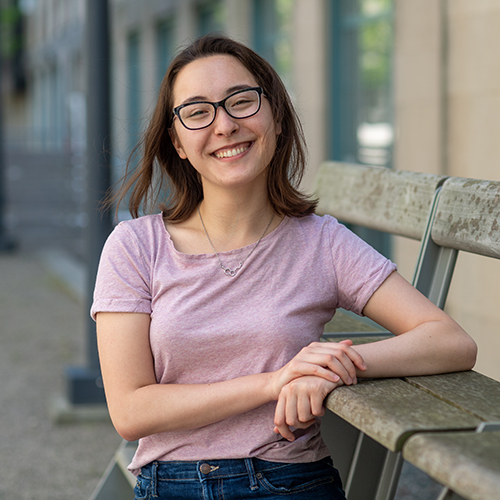 Photo of Newbury center grad Maelee Chen. Chen wears a pink tee shirt and jean shorts as she poses next to a wooden bench/ she sits next to it as her right elbow rests on the seat, and smiles to the camera.