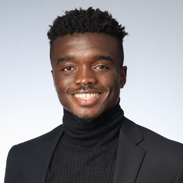 Photo of Peter Kargbo (SHA’22). Kargbo, a young black man with a short afro haircut and wearing a black turtleneck and blazer smiles for the camera.
